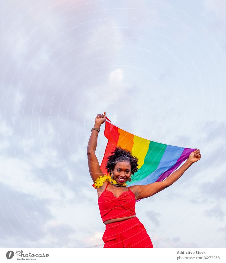 Lächelnde schwarze Frau mit LGBTQ-Flagge auf der Straße Fahne stylisch Mode feiern Individualität Stolz Spektrum anheben Arme hochgezogen lgbtq Stil