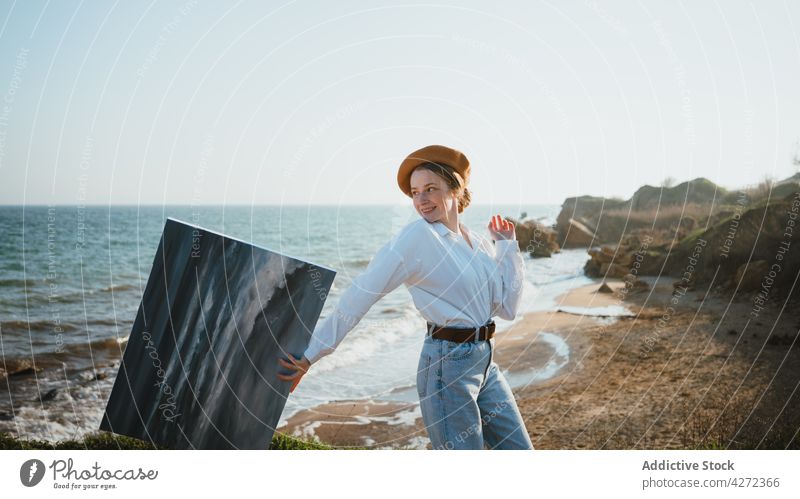 Lächelnde junge Frau, die mit einem Gemälde in der Hand am Meer spazieren geht Spaziergang Meeresufer Malerei Natur Inhalt Glück Anstreicher Strand Stil positiv