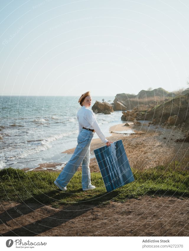 Junge Frau, die mit einem Gemälde in der Hand am Meer spazieren geht Spaziergang Meeresufer Malerei Natur Anstreicher Strand Stil MEER Küste jung trendy Hut
