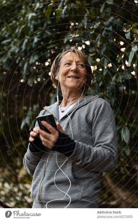Positive ältere Frau, die während des Trainings Musik auf ihrem Smartphone hört gealtert zuhören benutzend Fitness Kopfhörer positiv Sportbekleidung Gerät