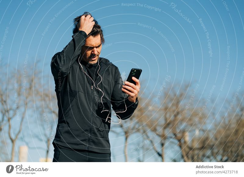 Sportler mit Kopfhörern beim Surfen auf dem Smartphone im Stadtpark plaudernd achtsam zuhören Musik maskulin Park benutzend Apparatur Mann Großstadt Gerät Fokus