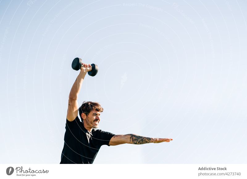 Tätowierter Sportler beim Training mit Hantel im Sonnenlicht Arme hochgezogen Kurzhantel Ausdauer Motivation Tattoo Spannung Mann Gewicht anheben Himmel