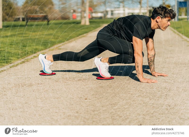 Entschlossener Sportler, der im Park mit Übungsscheiben trainiert Athlet Plankenhaltung Ausdauer Training anstrengen Mann Straßenbelag Motivation Tattoo