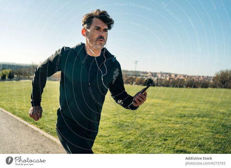Aktiver Läufer joggt auf dem Gehweg im Stadtpark joggen Sport Aktivität Training Herz Smartphone Kopfhörer Musik Mobile schnell Mann Park Energie Jogger laufen