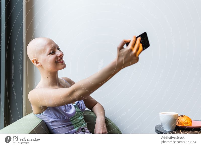 Glatzköpfige Frau macht Selfie mit Smartphone auf Couch in der Nähe von Tisch mit Tasse unter Sofa kahl trinken Raum modern sitzen benutzend Alopezie Apparatur