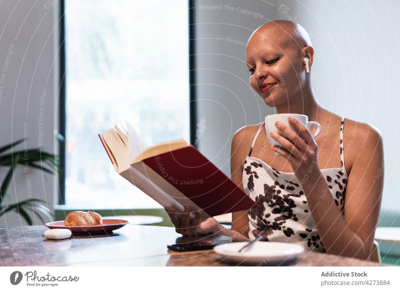 Positive junge haarlose Frau trinkt Kaffee und liest einen Roman in einer Cafeteria lesen Buch trinken Café Alopezie sich[Akk] entspannen Lächeln genießen