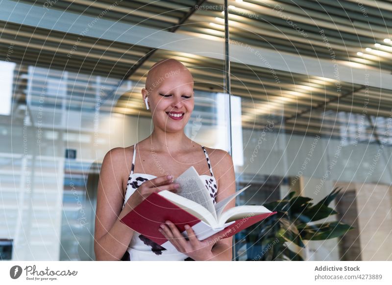 Lächelnde junge haarlose Dame, die am Arbeitsplatz ein Buch liest Frau lesen Alopezie Glück positiv Etage Büro Stil Persönlichkeit kahl trendy tws Kopfhörer
