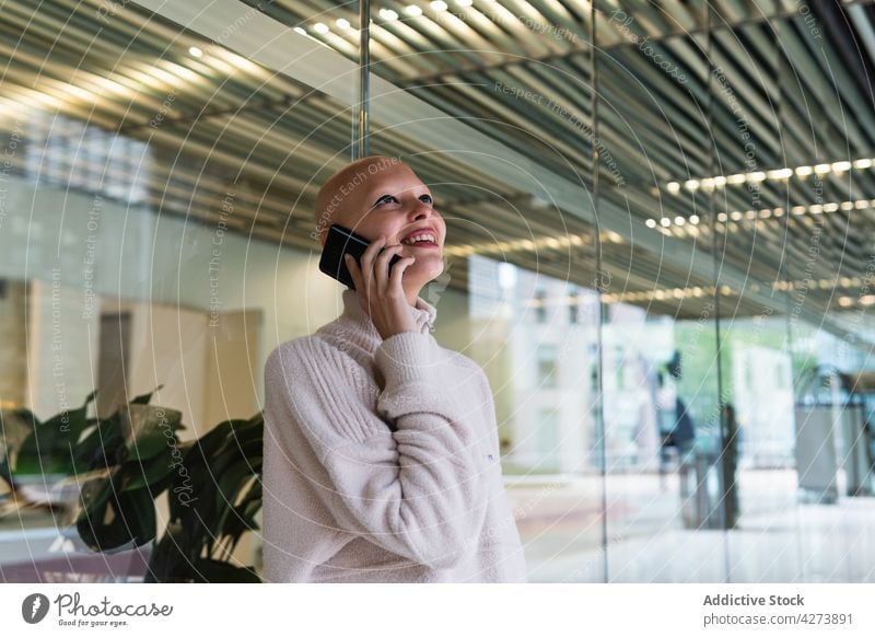 Lächelnde junge Frau mit Glatze, die im Büro mit ihrem Smartphone spricht reden Telefonanruf Alopezie Gespräch Kommunizieren diskutieren positiv Arbeit kahl