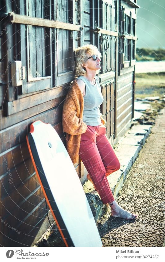 Porträt einer jungen reifen blonden kaukasischen Frau im Freien in einem Holzhaus in der Nähe eines Strandbereichs mit einem Bodyboard und einer Sonnenbrille.  Lifestyle-Konzept.