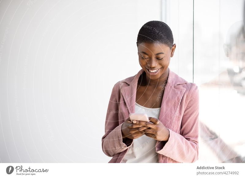 Fröhliche schwarze Frau, die ihr Smartphone gegen eine Glaswand hält trendy Mode heiter Browsen nachdenken Nachricht offen benutzend Apparatur Gerät Funktelefon