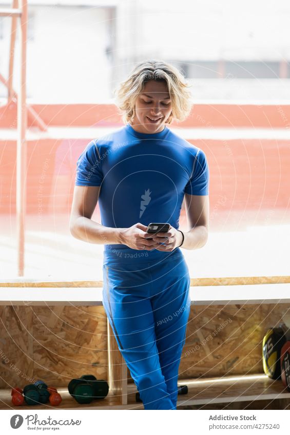 Lächelnder Sportler, der im Fitnessstudio mit seinem Smartphone chattet plaudernd Internet online Beine gekreuzt Freizeit benutzend Apparatur Mann Gerät