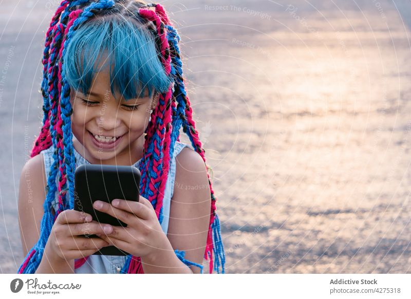 Lächelndes ethnisches Mädchen, das im Sonnenlicht eine Textnachricht auf einem Smartphone schreibt Internet Surfen online heiter Zeitvertreib Kindheit Geflecht