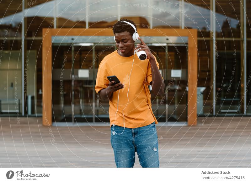 Junger schwarzer Mann genießt Musik mit Kopfhörern meloman Lächeln zuhören benutzend Smartphone Gesang Freizeit Wiedergabeliste Browsen männlich Gebäude Glück