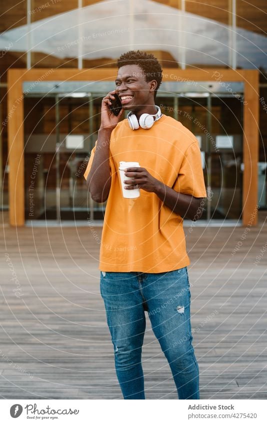 Fröhlicher schwarzer Mann mit einer Tasse Kaffee, der mit seinem Smartphone spricht sprechen Lachen benutzend Kopfhörer zum Mitnehmen Getränk reden Gespräch