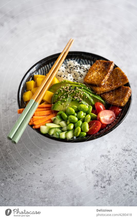 Seitenansicht einer hausgemachten veganen Poke Bowl mit Tofu, Mango, Avocado, Karotte, Edamame, Tomate, Gurke auf grauem Hintergrund, vertikal asiatisch