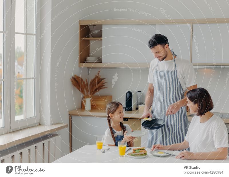 Vater bereitete Spiegeleier für Familie, kleines Kind hält Teller und wartet auf das Frühstück. Familie posieren in der Küche in der Nähe von Tisch, genießen Sie leckere Mahlzeit, haben frohe Ausdrücke. Menschen, Essen, häusliche Atmosphäre