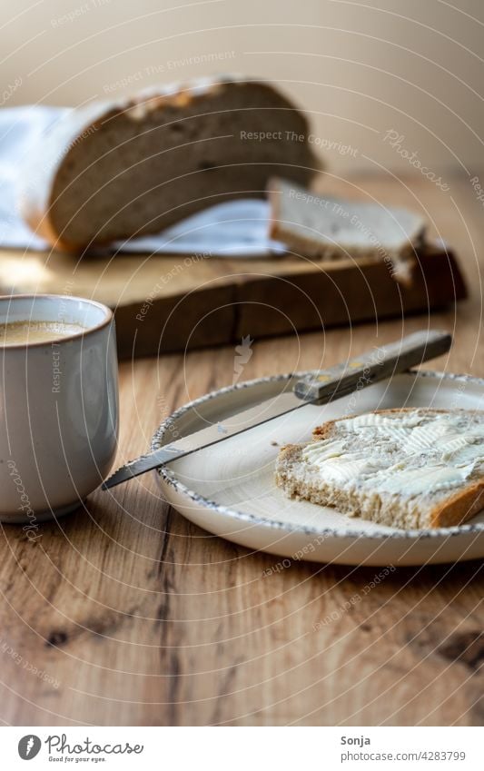 Frühstückstisch mit einer Tasse Kaffee und einer Scheibe Brot mit Butter Brotscheibe Kaffeetasse Teller Holztisch rustikal Messer Morgen Nahaufnahme