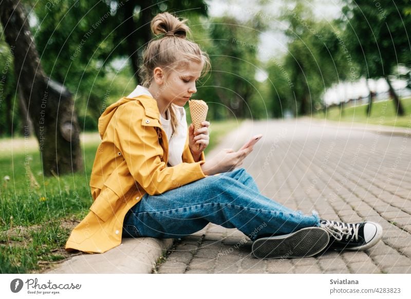 Ein blondes Mädchen sitzt mit einem Telefon und Eis auf der Straße und kommuniziert in sozialen Netzwerken. Kind im Freien Sommer Park Teenager Smartphone