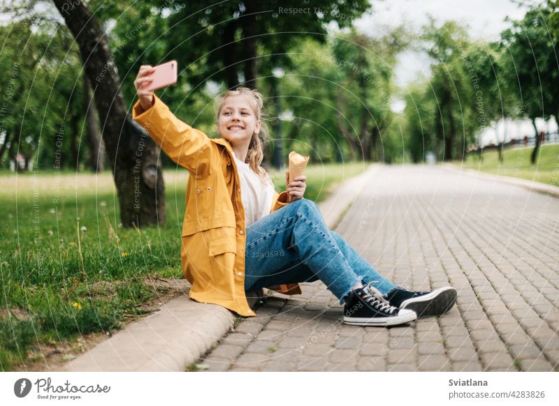 Eine Blondine sitzt auf der Straße mit einem Telefon und Eis macht ein lustiges Selfie. Kind im Freien Sommer Park Teenager Smartphone Speiseeis