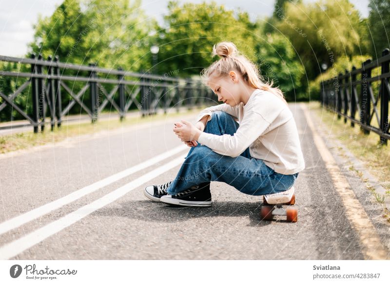 Nettes blondes Teenager-Mädchen, das auf einem Skateboard in einem Stadtpark sitzt, mit Freunden auf einem Smartphone chattet und ein Selfie macht.