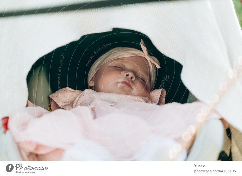Ein kleines süßes Baby-Mädchen in einem rosa Kleid schläft süß im Kinderwagen. Nahaufnahme. Weicher Fokus. Ein schöner Säugling ruht, während er im Park spazieren geht.Glückliche Kindheit Konzept. Lebensstil Fotografie.