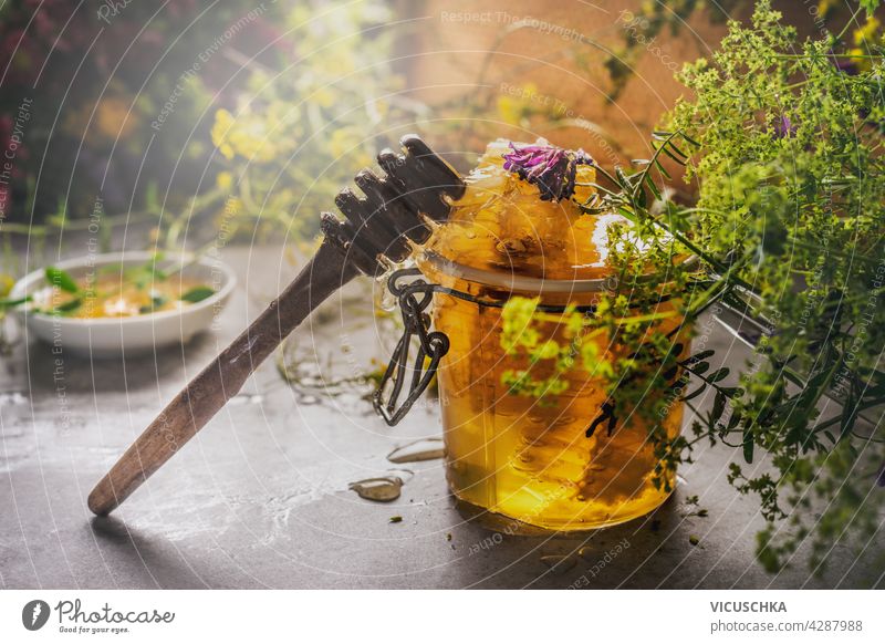Nahaufnahme eines Glases mit Honig und Honiglöffel und Wildblumen. Schale mit Honig im Hintergrund. Natürliche Lebensmittel Konzept. Dunkler Beton-Hintergrund. Vorderansicht.