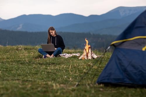 junge weibliche Freiberufler arbeiten am Laptop in den Bergen am Abend. Tourist Mädchen sitzt in der Nähe von Lagerfeuer und Zelt. Kopierraum Sitzen Frau Urlaub