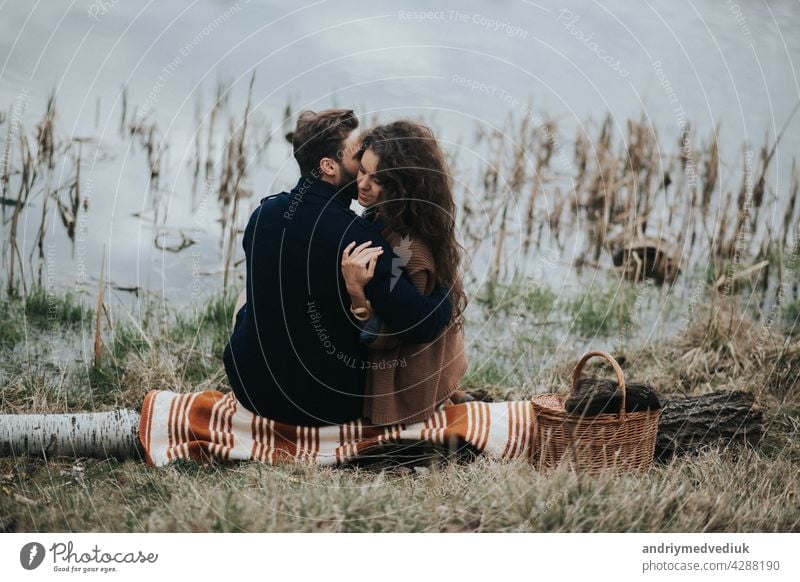 zwei kaukasische Liebhaber sitzen auf Decke am See. Junges Paar umarmt sich am Herbsttag im Freien. Ein bärtiger Mann und lockige Frau in der Liebe. Valentinstag. Konzept der Liebe und Familie. Rückansicht