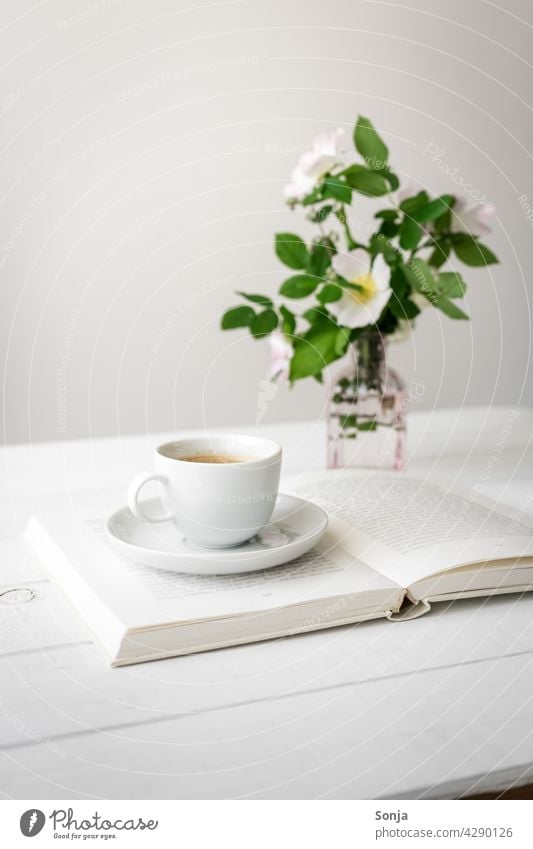 Eine Tasse Kaffee auf einem offenen Buch und Blumen in einer Vase auf einem weißen Holztisch Bucht Stillleben zuhause Kaffeepause Frühstück Kaffeetrinken