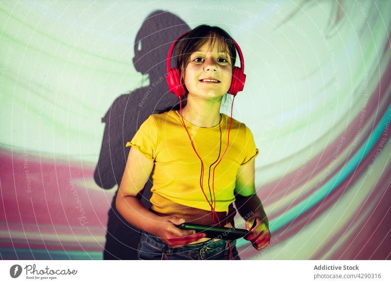 Zufriedenes Mädchen, das ein Tablet benutzt und über Kopfhörer Musik hört zuhören Tablette Browsen Apparatur heiter Inhalt neonfarbig Licht Kind lässig Surfen
