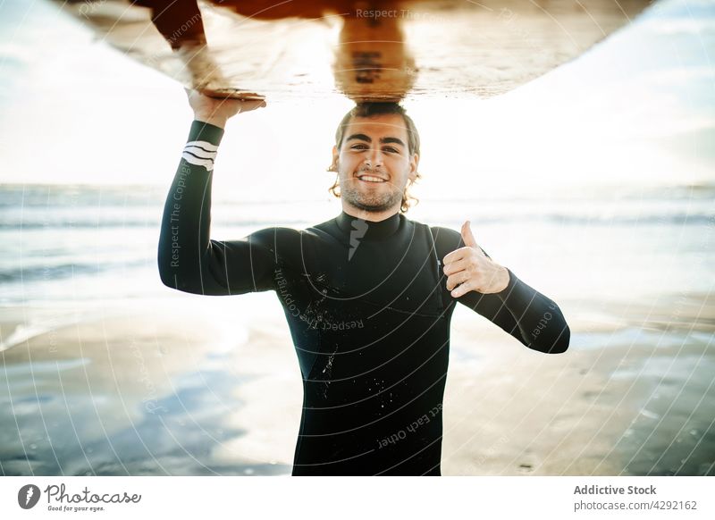 Lächelnder Surfer am Strand mit Surfbrett Mann Natur Sonnenuntergang Porträt winken im Freien Neoprenanzug Meeresküste männlich Daumen hoch Brandung Glück cool