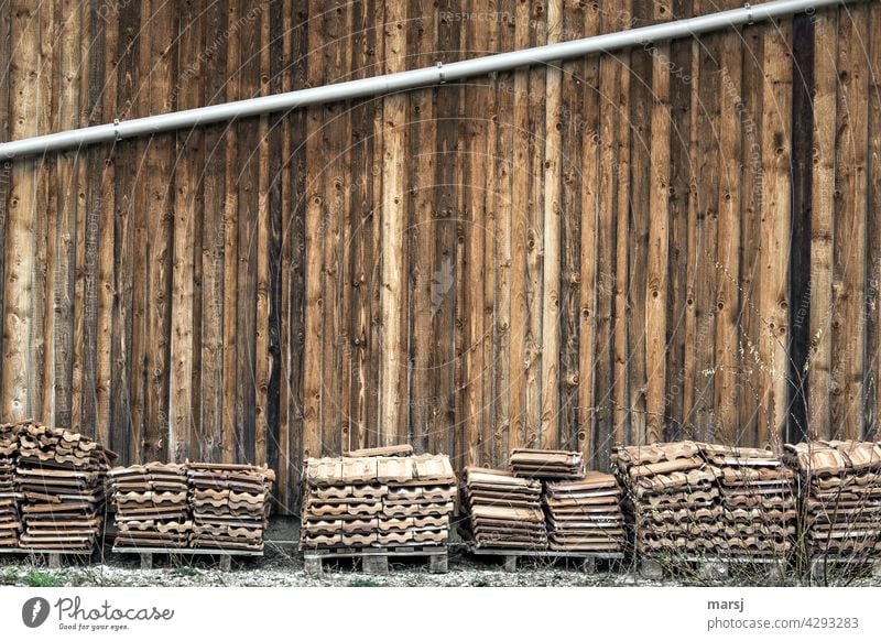 Schräg verlaufendes Fallrohr an einer Holzfassade mit verschiedenen Palletten mit Ziegeln. Dachziegel gelagert Paletten einzigartig Ordnungsliebe einfach braun