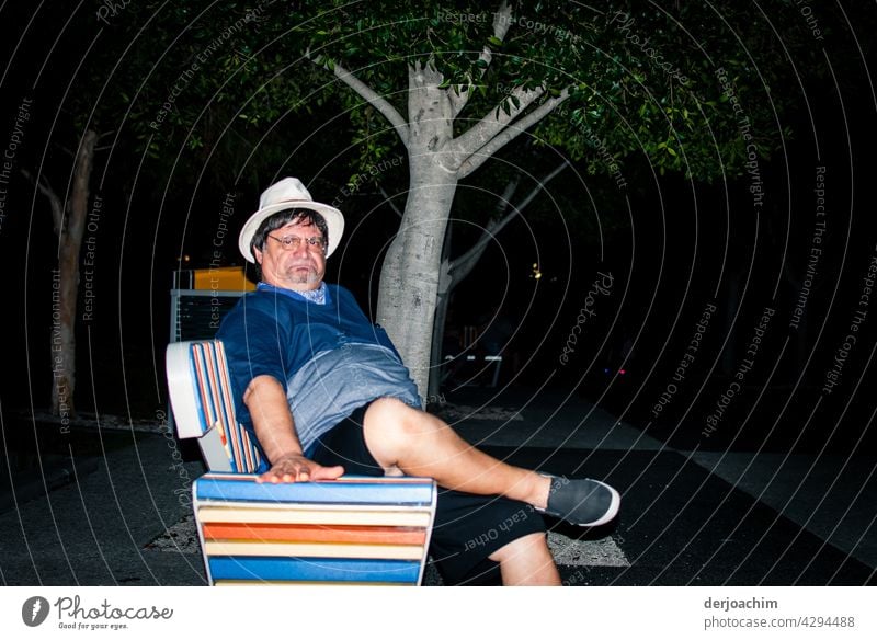 " Er ist  sauer " Mann im mittleren Alter, sitzt auf einer bunten Parkbank am Abend lässig  mit weißem Hut und Brille , kurzen Hosen und Sandalen und verzieht das Gesicht.