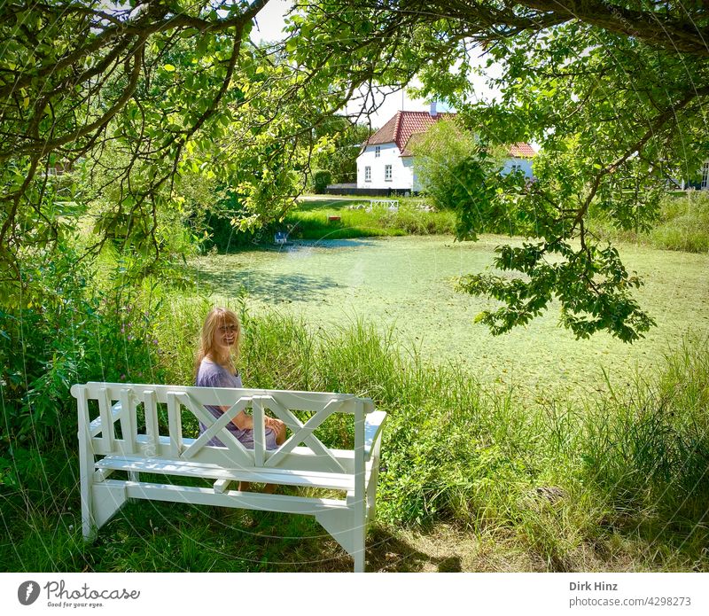 Attraktive blonde Frau sitzt auf weißer Bank unter einer Eiche an einem Teich schön feminin Erwachsene Außenaufnahme Sitzbank Mensch Idylle Dänemark Sommer