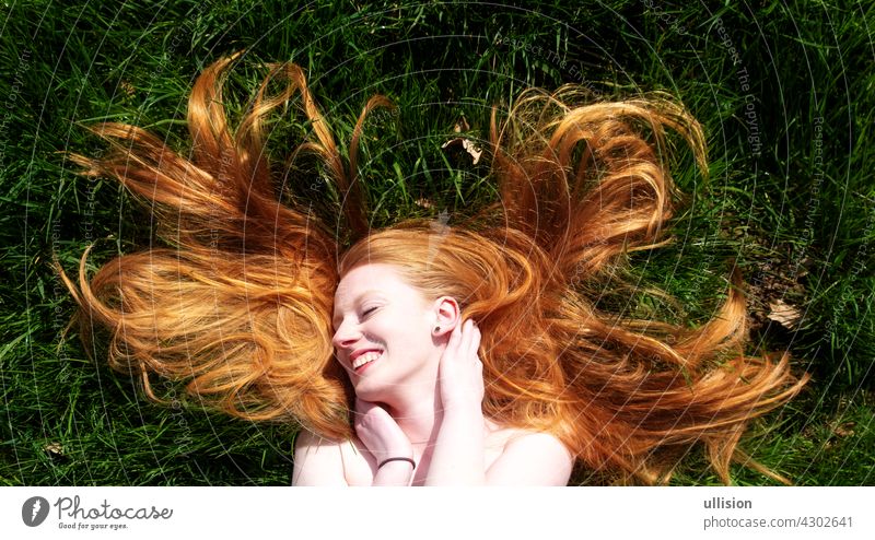 Schönes Porträt einer jungen sexy rothaarigen Frau, die glücklich in der Sommersonne liegt, lacht und sich auf dem grünen Gras entspannt, die roten Haare frei um den Kopf drapiert. Kopierraum