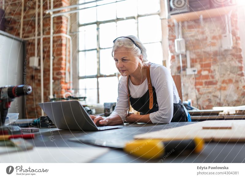 Ältere Frau mit Laptop in ihrer Werkstatt im Innenbereich Mechaniker Besitzer Beruf Dienst Kleinunternehmen Mitarbeiter arbeiten Techniker Arbeitsplatz Arbeiter