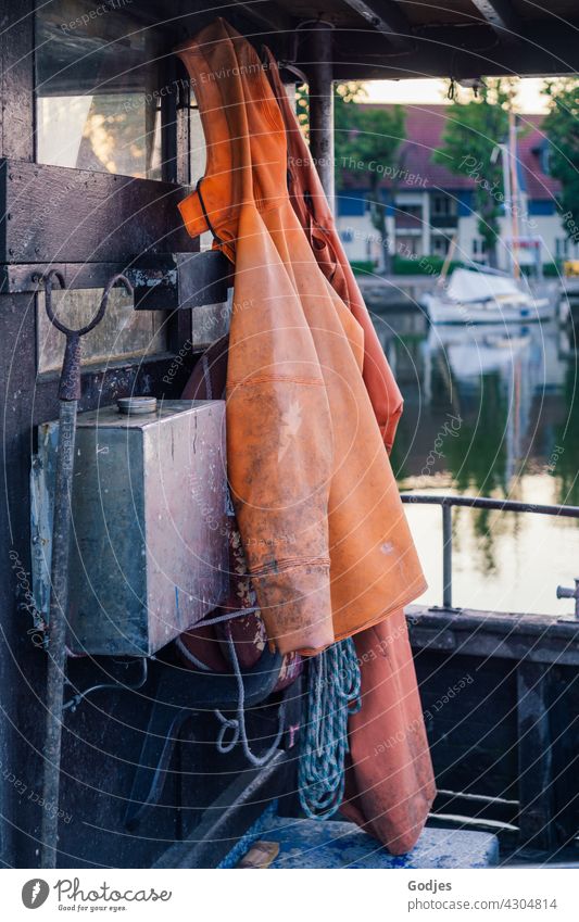 Friesennerz, Hacke, Seil hängen auf einem Fischerboot. Im Hintergrund ein Segelboot auf dem Wasser und ein Haus Regenmantel Regenhose Bekleidung friesennerz