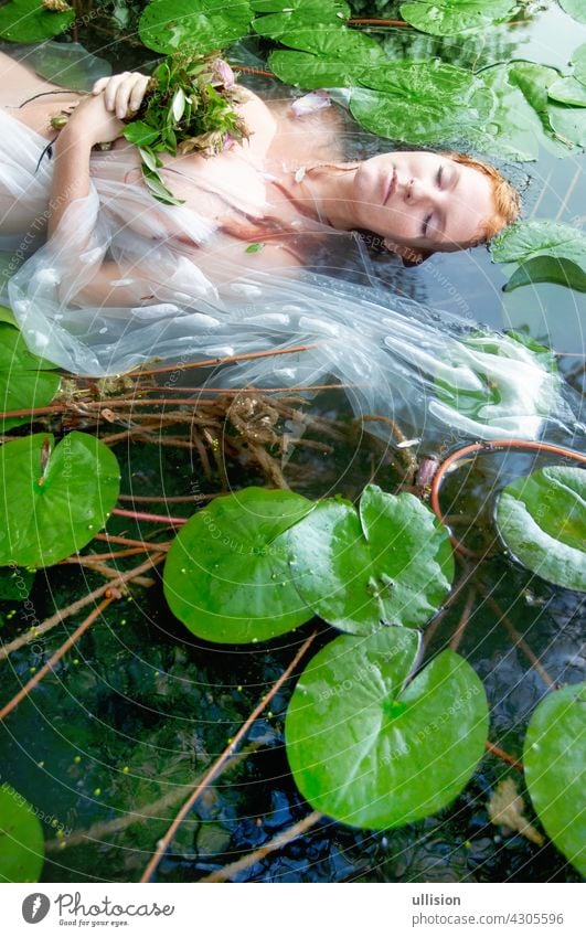Junge sexy Frau Ophelia mit lockigen roten Haaren liegt mit den Blumen mit geschlossenen Augen im weißen Kleid sterben im Wasser im See Braut ophelia Schönheit