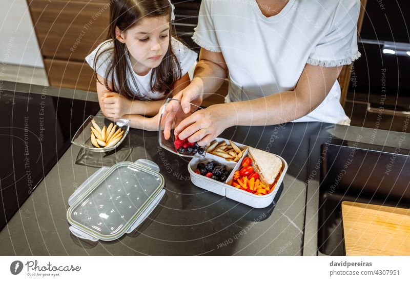 Mädchen beobachtet ihre Mutter setzen Essen in ihre Lunch-Box Setzen Frucht organisch Tochter Blick Schule gesunder Snack Veganismus vorbereitend Brotstangen