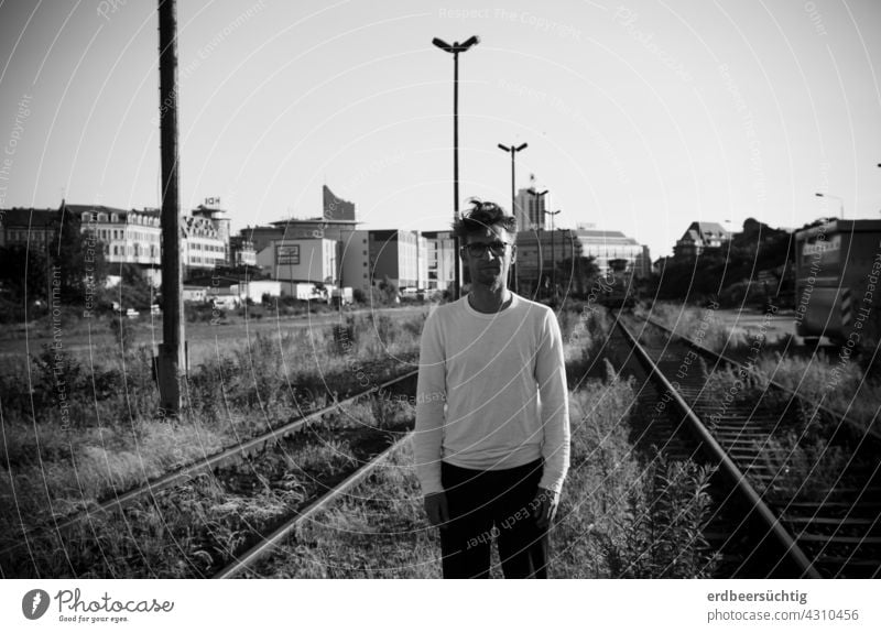 Mann steht etwas verloren zwischen stillgelegten Gleisen vor Stadtkulisse schwarz-weiß BRACHE zugewuchert Pflanzen Eisenbahn lost lost places Stadtsilhouette