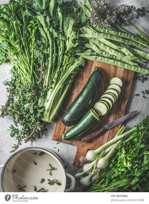Grünes gesundes Gemüse und Kräuter Zutaten für schmackhafte vegane Küche auf Schneidebrett und rustikalen Küchentisch mit leeren Kochtopf. Ansicht von oben grün