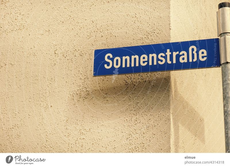 blaues Straßennamenschild mit weißer Schrift " Sonnenstraße " an einer Metallstange vor einer Hausecke mit Schatten / wohnen / Navigation Straßenschild