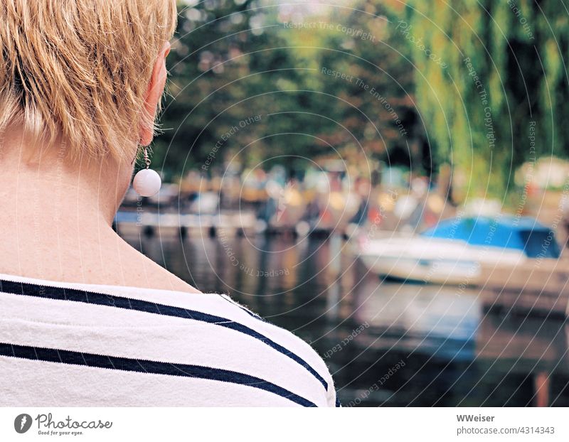 Eine Frau mit Kurzhaarschnitt und modischem Ohrring schaut hinüber zu einem Bootshafen mit vielen Leuten Dame Rücken Sommer modern schick Streifen maritim