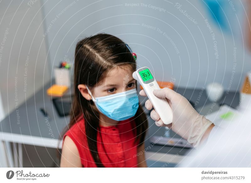 Mädchen ohne Fieber, dessen Temperatur vom Arzt gemessen wird Infrarot-Thermometer kein Fieber Schutzmaske Messung ärztliche Untersuchung Stirn grün Coronavirus