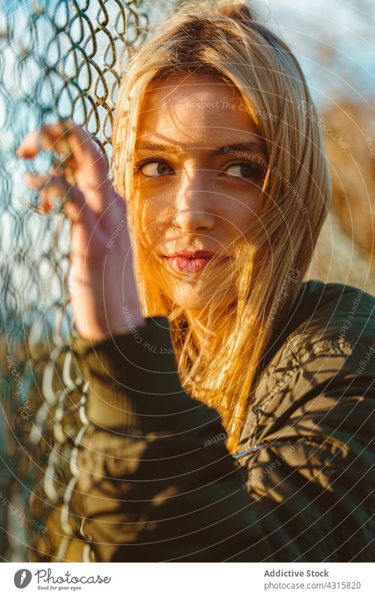 Ruhige Frau lehnt sich im Sonnenlicht an einen Netzzaun Zaun Porträt Kettenglied Windstille blond charmant modern Schönheit jung hell sorgenfrei allein Jacke