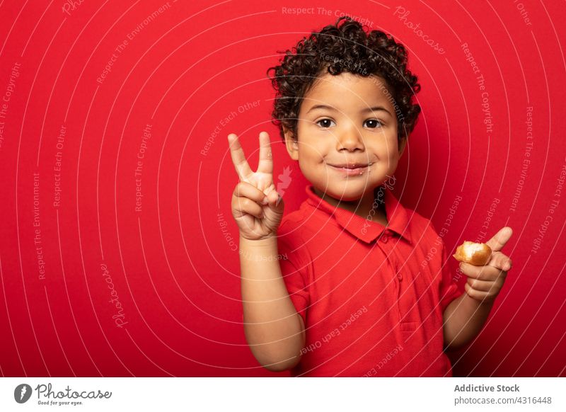 Zufriedener Junge mit Donut und Zwei-Finger-Geste Kind v-Zeichen zwei Finger gestikulieren essen Doughnut Krapfen Sieg geschmackvoll lecker süß Kindheit heiter