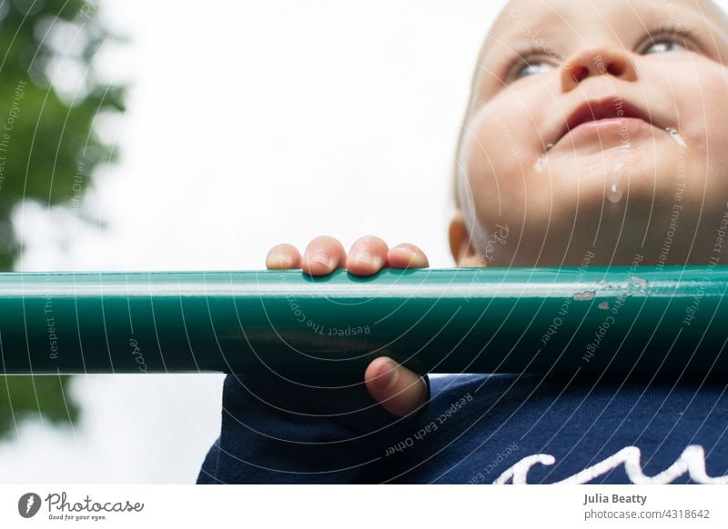 Beißendes Kleinkind auf dem Spielplatz; Baby zieht sich zum Stehen hoch, indem es das grüne Metallbecken auf dem Gerät benutzt zahnend sabbernd Zähne hochziehen