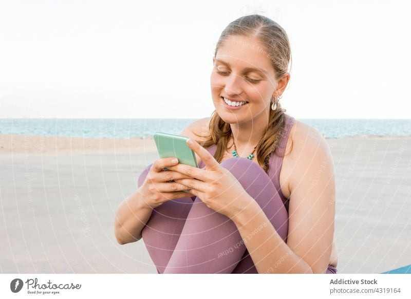Lächelnde Frau mit Smartphone auf der Yogamatte Mobile Telefon Yogi Strand Apparatur Browsen heiter benutzend Glück schlank Textnachricht Gerät sitzen Unterlage