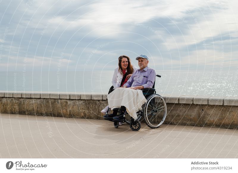 Fröhliche erwachsene Tochter und betagter Vater im Rollstuhl auf der Promenade Mann Senior Kälte Spazierweg MEER Stauanlage Zusammensein Meeresufer Erwachsener