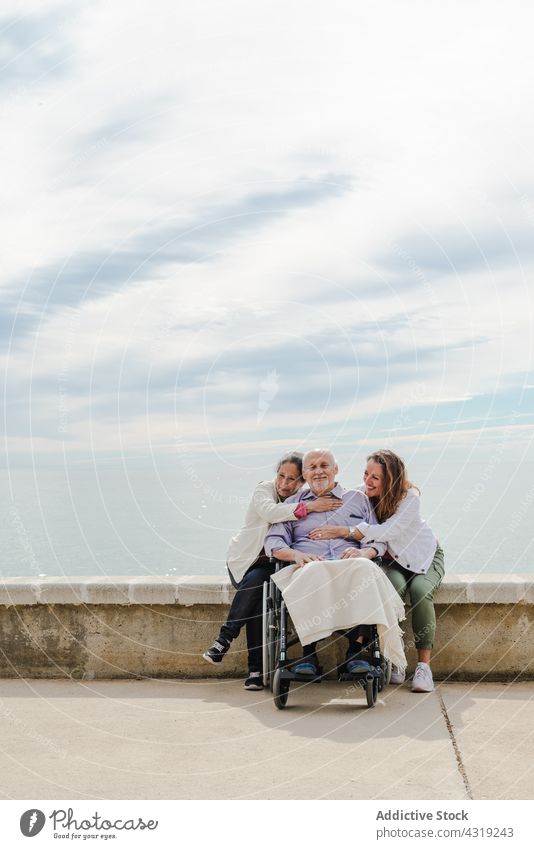 Fröhliche Familie umarmt Vater im Rollstuhl Senior Spazierweg Umarmung Einheit Kälte Stauanlage Zusammensein MEER Meeresufer Erwachsener deaktivieren Handicap